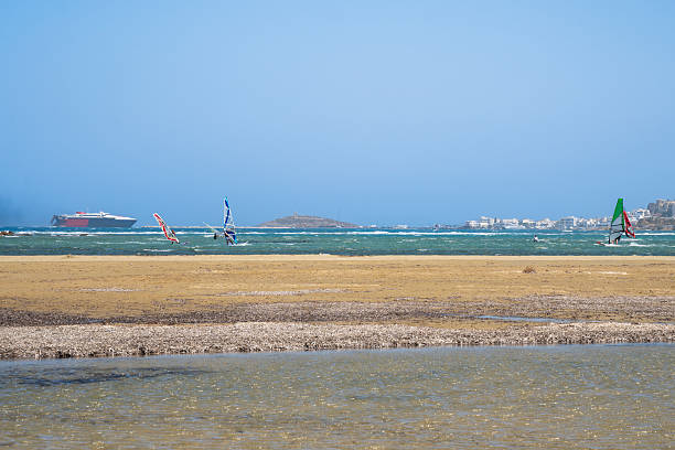 Windsurfers glide across a shallow lagoon with waves and a distant ferry on the horizon. Captured near Naxos, Greece, this scene highlights dynamic watersports and clear Mediterranean summer conditions.
