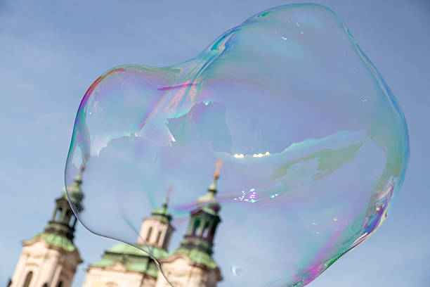 Giant soap bubble catching iridescent colors and reflecting the historic saint nicholas church spires under a bright blue sky, creating a whimsical and playful scene in prague, czech republic