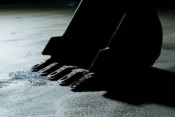 Steel excavator bucket teeth positioned on a clean ground surface under strong lighting, representing essential heavy equipment used in industrial earthmoving, construction workflow