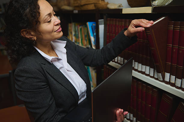 istock Professional Woman Browsing Library Bookshelf with Laptop in Hand 2248922347