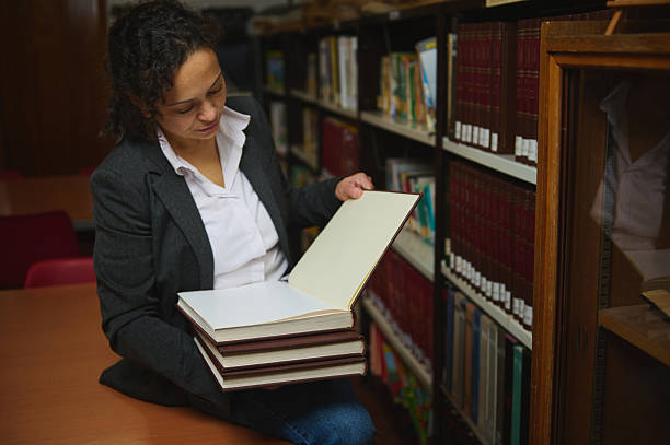 istock Professional Woman In Library Reviewing Open Notebook While Stacks Of Books Rest In Her Lap 2248923185