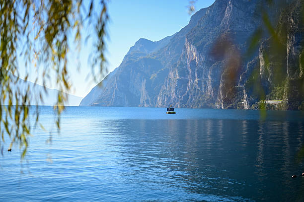 View of a wooden pier and small beach on Lake Garda, framed by a tree and lush greenery near Brenzone sul Garda, Veneto region, Italy. Mountains in the background.