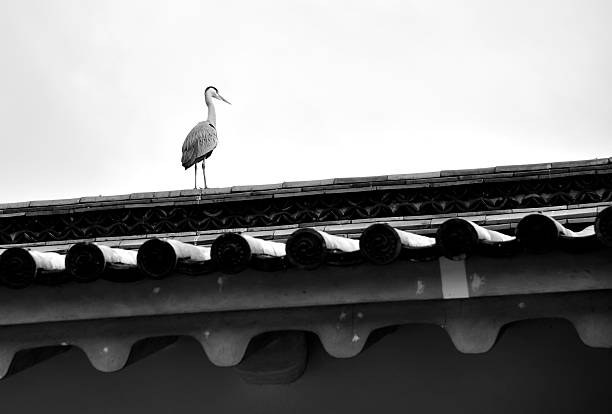 A heron standing on a traditional Japanese tiled roof against a cloudy sky.