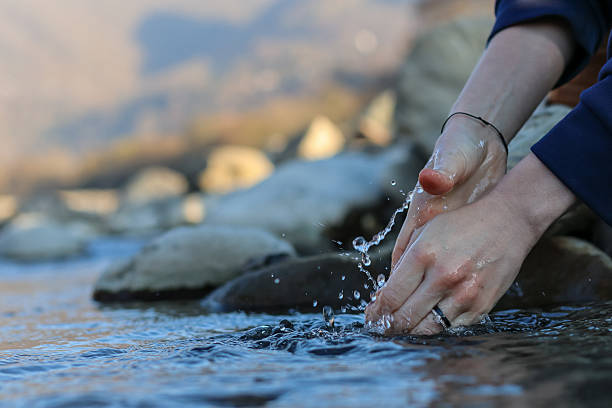 A female hand wearing a thin bracelet touches shallow stream water, sending ripples across rocks in a serene outdoor setting.