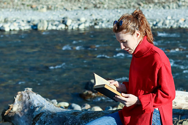 Young woman reading book near lake on sunny day