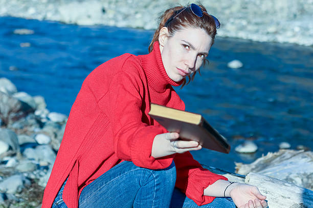 Woman in red sweater sits on rocks near river, extending closed book forward with stern look.