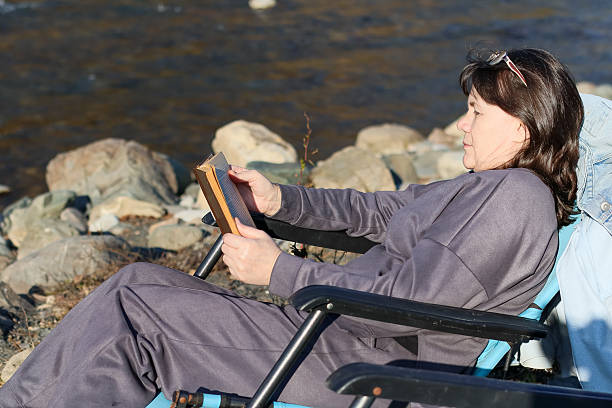 Close-up side profile of woman in casual attire reading a novel outdoors on a chair beside a stony river shore