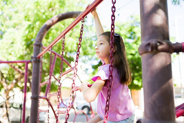 Girl holding onto playground bars, climbing the structure outdoors. Child enjoying playtime and recreation in a bright park setting