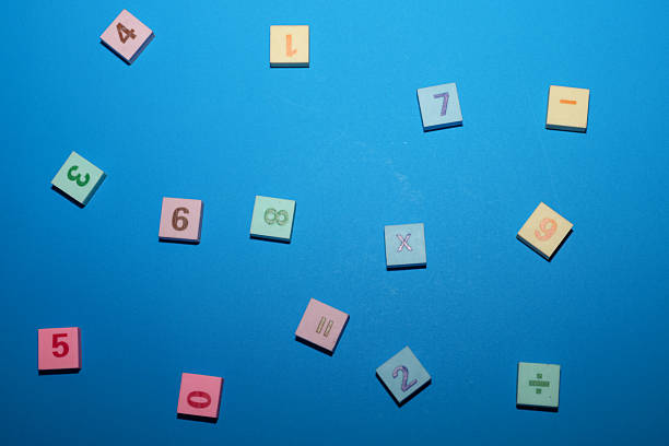 Small square rubber tiles or erasers with various numbers and mathematical symbols are randomly scattered across a bright blue background. The tiles feature soft pastel shades, including pink, yellow, and light green.