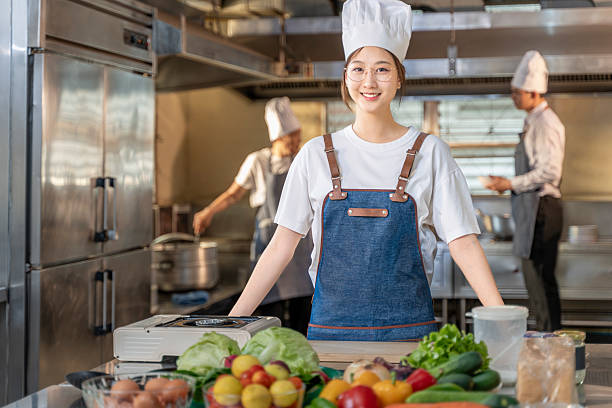 istock Happy young Asian female chef standing in her kitchen confidently smiling at camera while two other chefs working in the background 2248975279