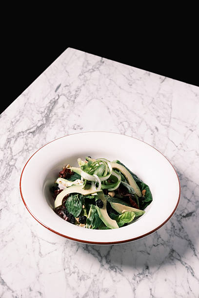High angle view of a warm Caesar salad with shaved avocado, leafy greens, dried berries and parmesan flakes in a deep white bowl, Contemporary Italian restaurant interpretation of a classic dish, Culinary concept for menu design and modern food styling