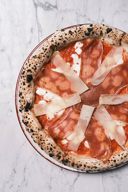 Top view of Neapolitan-style prosciutto crudo pizza with tomato sauce, melted cheese and thin cured ham slices on charred crust over marble table, Classic Italian restaurant specialty, Culinary concept for menu layouts and gourmet branding