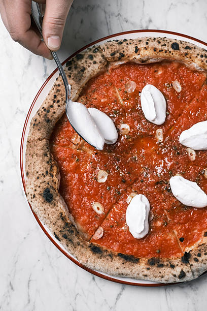 Top view of Neapolitan-style marinara pizza topped with garlic slices and added ricotta dollops being spooned on charred crust over marble table, Classic Italian restaurant preparation moment, Culinary concept for menu design and artisan food branding