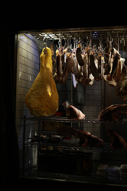 Close-up view of assorted cured meats and large aged ham hanging on metal hooks inside a refrigerated storage unit in a restaurant kitchen, Warm focused lighting highlighting texture and preservation detail, Professional butchery and culinary preparation