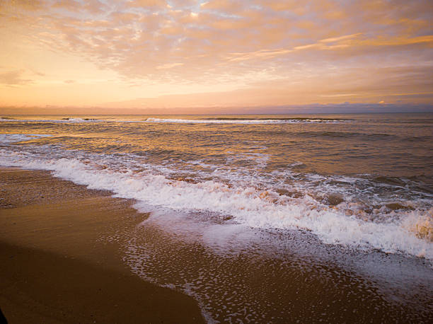 A beautiful, wide-angle shot of the ocean during the golden hour. The sky is filled with soft orange and pink hues, reflecting off the water. Waves crash onto the dark, wet sand, creating white foam.