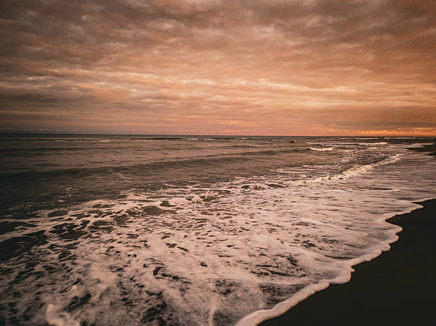 A dramatic, moody coastal scene at sunset or dusk. Dark, turbulent ocean waves wash onto a black or dark sand beach, creating thick white seafoam. The expansive sky is filled with warm, golden-orange and brown clouds, giving the image a nostalgic, cinematic feel.