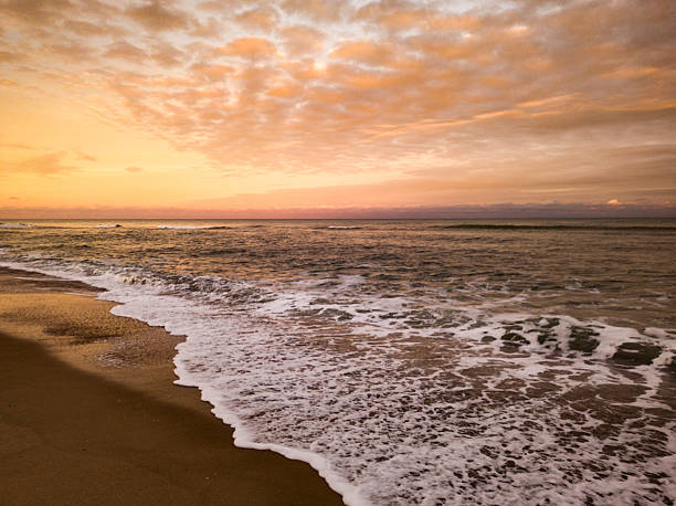 A dramatic view of the ocean shoreline during golden hour. The sky is filled with warm orange and peach clouds above the dark horizon. Waves crash gently onto the wet, dark sand, creating a line of white foam.