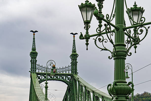Budapest, Hungary - April 2025 The iconic Elizabeth Bridge, a historic bridge painted in green, stretches across the Danube River in Budapest, Hungary. Featuring intricate ironwork, tall gothic-style towers topped with statues, and vintage street lamps along its railings, the structure connects the Buda and Pest sides of the city. The river flows calmly below under an overcast sky, with a few distant pedestrians visible on the bridge. This image captures the architectural grandeur and engineering marvel of the 19th-century landmark on a cloudy daytime in autumn or spring. Ideal for commercial use in travel magazines, tourism campaigns, historical documentaries, urban planning articles, and European architecture features, highlighting Budapest's skyline and riverfront heritage.