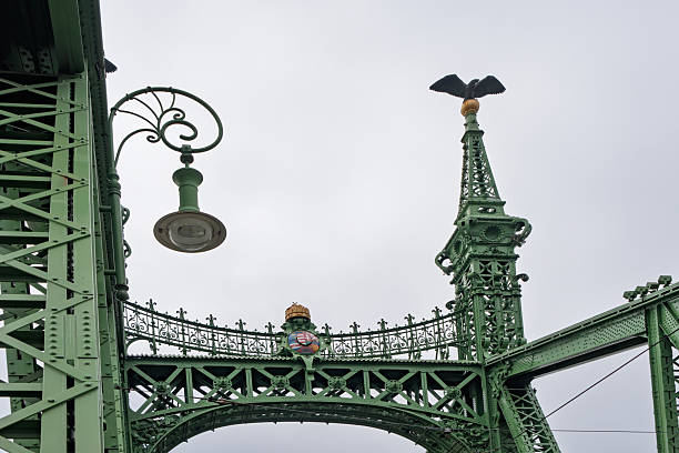 Budapest, Hungary - April 2025 The iconic Elizabeth Bridge, a historic bridge painted in green, stretches across the Danube River in Budapest, Hungary. Featuring intricate ironwork, tall gothic-style towers topped with statues, and vintage street lamps along its railings, the structure connects the Buda and Pest sides of the city. The river flows calmly below under an overcast sky, with a few distant pedestrians visible on the bridge. This image captures the architectural grandeur and engineering marvel of the 19th-century landmark on a cloudy daytime in autumn or spring. Ideal for commercial use in travel magazines, tourism campaigns, historical documentaries, urban planning articles, and European architecture features, highlighting Budapest's skyline and riverfront heritage.