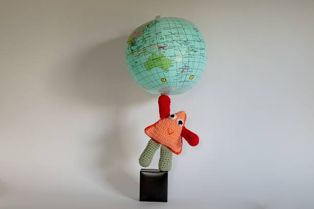 A soft toy fantasy character stands on one leg of top of a black box, balancing a globe on one hand