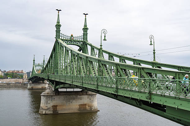 Budapest, Hungary - April 2025 The iconic Elizabeth Bridge, a historic bridge painted in green, stretches across the Danube River in Budapest, Hungary. Featuring intricate ironwork, tall gothic-style towers topped with statues, and vintage street lamps along its railings, the structure connects the Buda and Pest sides of the city. The river flows calmly below under an overcast sky, with a few distant pedestrians visible on the bridge. This image captures the architectural grandeur and engineering marvel of the 19th-century landmark on a cloudy daytime in autumn or spring. Ideal for commercial use in travel magazines, tourism campaigns, historical documentaries, urban planning articles, and European architecture features, highlighting Budapest's skyline and riverfront heritage.