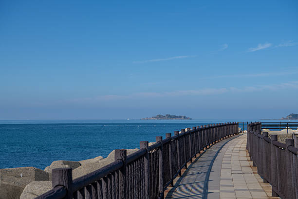 Photograph of Hashima Island (Gunkanjima) viewed from Takahama Beach in Nagasaki, Japan.A curved coastal promenade leads the viewer’s eye toward the distant abandoned island, surrounded by calm blue sea and clear sky.Suitable for tourism promotion, historical context visuals, editorial features, travel media, and scenic background usage.