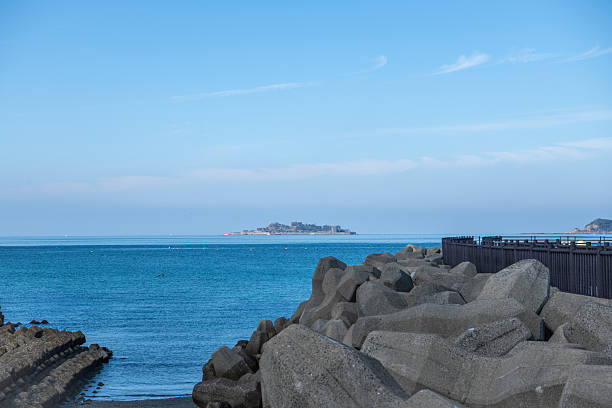 Photograph of Hashima Island (Gunkanjima) viewed from Takahama Beach in Nagasaki, Japan.A curved coastal promenade leads the viewer’s eye toward the distant abandoned island, surrounded by calm blue sea and clear sky.Suitable for tourism promotion, historical context visuals, editorial features, travel media, and scenic background usage.