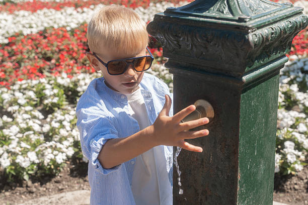 A young boy presses a metal well faucet with his hand while standing in a bright garden surrounded by blooming flowers. The close-up captures movement, curiosity, and the warm atmosphere of a sunny summer day.
