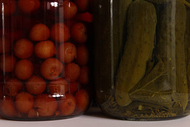Macro crop showing red fruit compote beside packed pickled cucumbers through the glass. Rich colors and textures of homemade preserves.
