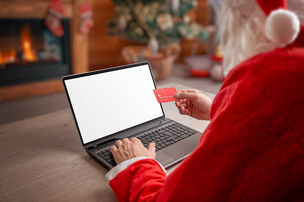 istock Santa using laptop with blank screen while holding credit card in cozy Christmas interior, perfect for holiday mockups, ecommerce concepts and festive digital designs 2249008009