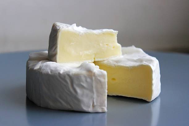 Close-up of a soft brie or camembert cheese wheel with creamy wedges stacked on top. White edible rind, smooth interior, neutral background, copy space.
