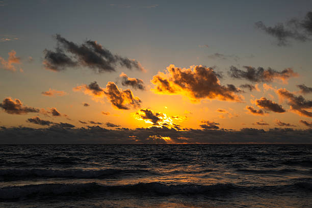 An atmospheric sunrise paints the horizon in warm tones at Lauderdale-by-the-Sea. Waves gently blur with long exposure, while dark clouds create a dramatic contrast against the glowing sky. The sandy shoreline and scattered seaweed add natural texture, evoking a mood of tranquility, new beginnings, and the quiet power of nature. Ideal for themes such as mindfulness, coastal landscapes, travel inspiration, nature’s beauty, and peaceful beach mornings.