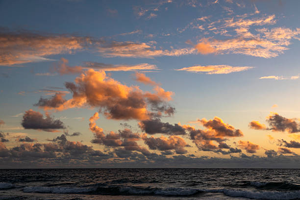 An atmospheric sunrise paints the horizon in warm tones at Lauderdale-by-the-Sea. Waves gently blur with long exposure, while dark clouds create a dramatic contrast against the glowing sky. The sandy shoreline and scattered seaweed add natural texture, evoking a mood of tranquility, new beginnings, and the quiet power of nature. Ideal for themes such as mindfulness, coastal landscapes, travel inspiration, nature’s beauty, and peaceful beach mornings.