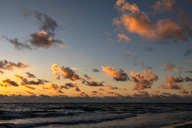 An atmospheric sunrise paints the horizon in warm tones at Lauderdale-by-the-Sea. Waves gently blur with long exposure, while dark clouds create a dramatic contrast against the glowing sky. The sandy shoreline and scattered seaweed add natural texture, evoking a mood of tranquility, new beginnings, and the quiet power of nature. Ideal for themes such as mindfulness, coastal landscapes, travel inspiration, nature’s beauty, and peaceful beach mornings.