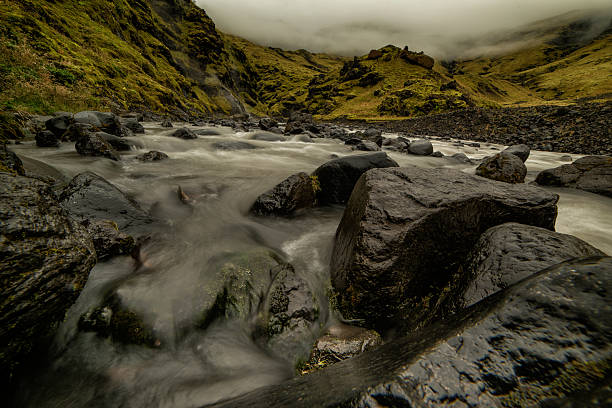 Icelandic landscape with a river flowing between green rocky hills under a moody, dramatic sky. Long-exposure shot with smooth, blurred water moving around dark stones, creating a peaceful yet powerful atmosphere.