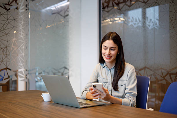 istock Joyful young professional woman focused on her mobile device while multitasking with a laptop in a modern shared workspace 2249020161