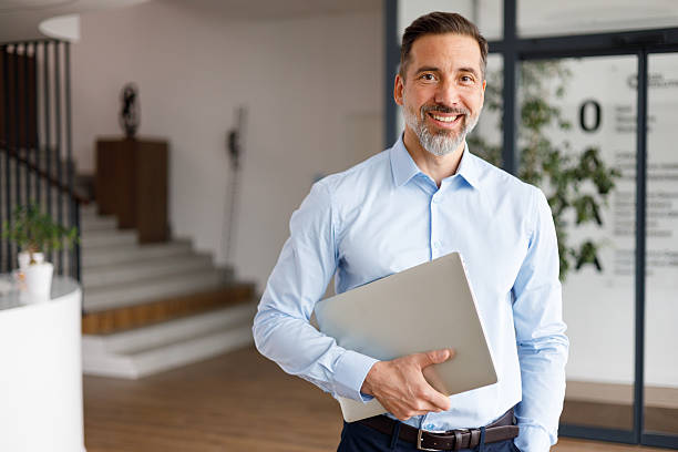 istock Businessman holding a laptop, smiling confidently while standing in a modern office environment, representing success and professionalism 2249022114