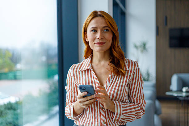 istock Professional woman with red hair using mobile phone in a contemporary office setting, texting, checking messages, smiling 2249024618