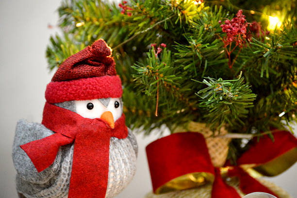 Cute plush bird toy wearing a red scarf and winter hat, positioned against a softly blurred Christmas tree background. The composition uses a shallow depth of field to focus sharply on the knitted fabric texture and bright red accessories, while the background hints at pine foliage and warm string lights, enhancing the winter holiday atmosphere. This charming visual is ideal for commercial use in marketing campaigns targeting seasonal cozy home decor, e-commerce banners for seasonal sales or gifts, social media content, and print media promoting traditional family celebrations.