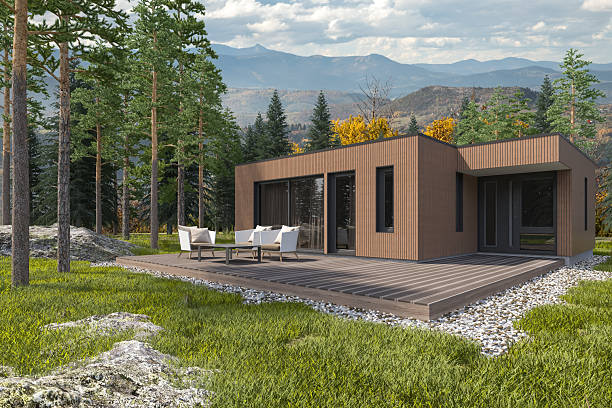 A contemporary weekend house featuring clean lines and minimalist Scandinavian architecture. The exterior is clad in vertical wooden panels, blending naturally with the forest surroundings. Large glass sliding doors open to a spacious wooden deck furnished with white outdoor chairs and a table. The cabin is nestled among pine trees, with mountains visible in the background. Gravel and rocks frame the deck, adding texture to the landscape. This peaceful retreat is ideal for showcasing modern rural living, sustainable architecture, or nature-inspired getaways. Suitable for use in real estate listings, travel blogs, or lifestyle editorials.
