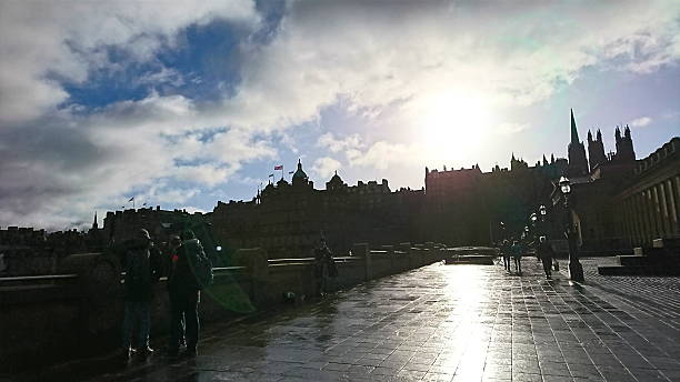 Edinburgh, UK - February 10 2018: People walk on a wet stone esplanade next to the The Royal Scottish Academy as the historic Edinburgh skyline is silhouetted against a dramatic sunlit sky with clouds