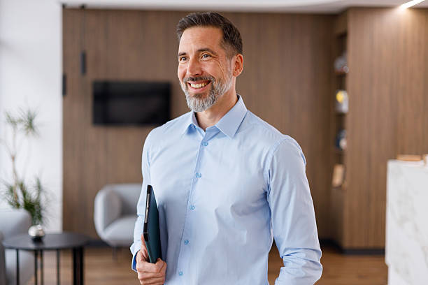 istock Entrepreneur standing in an office lobby, looking away and smiling. He is holding a folder, representing business and success 2249037548