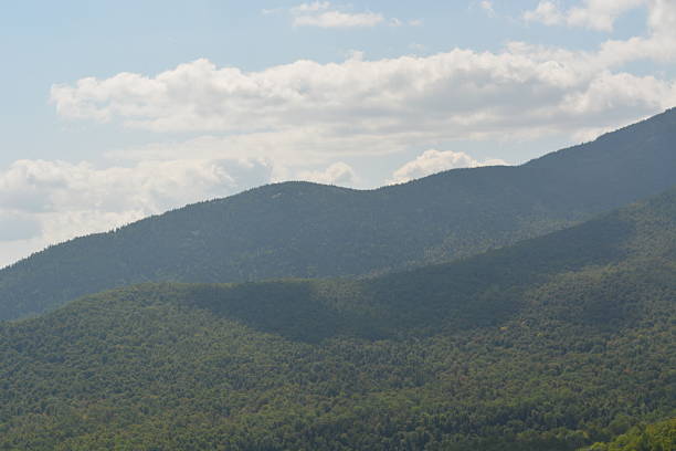 Vast green mountains rise majestically against a clear sky filled with fluffy clouds. Shadows play on the slopes, creating a serene and picturesque landscape during midday.