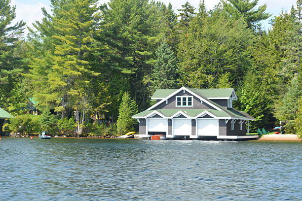A peaceful view of a stylish boathouse resting on the calm waters of a lake, framed by vibrant green trees. Kayaks and a dock add to the serene summer atmosphere.