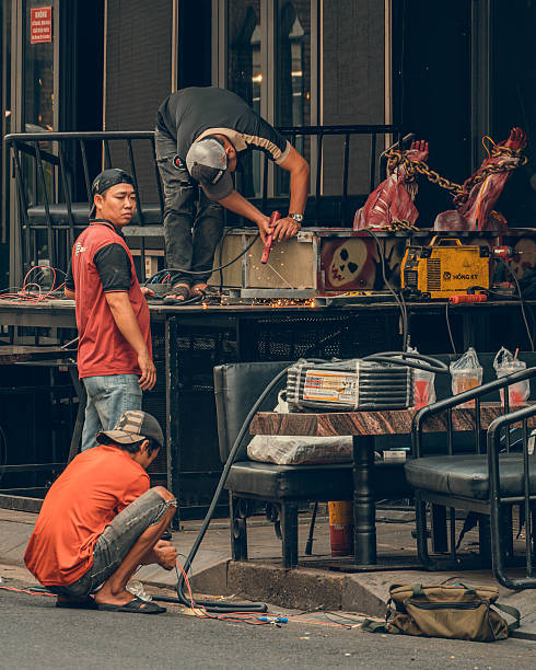 Ho Chi Minh City, Vietnam October 16, 2025 Three workers are busy with metal fabrication and electrical work on a sidewalk in a city. One man is welding or grinding metal on a black table, while two others are handling wires and equipment. A yellow HONG KY welding machine is visible. The scene captures a moment of industrious labor and craftsmanship in an outdoor setting.
