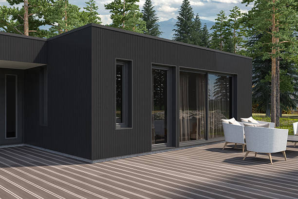 A modern countryside villa designed in Scandinavian style, featuring a sleek black pine wood facade. The rectangular structure includes large glass sliding doors that open to a wooden deck with white outdoor furniture. Surrounded by tall pine trees and mountain views, the house blends minimalist architecture with natural surroundings. Gravel and rocks frame the deck, adding texture and contrast to the dark exterior. The setting evokes a peaceful retreat ideal for weekend getaways or remote living. Perfect for showcasing sustainable design, rural architecture, or luxury cabin lifestyle. Suitable for real estate, travel editorials, or architectural portfolios.