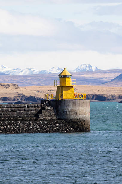 A bright yellow lighthouse stands on a rocky breakwater by the sea in Iceland.