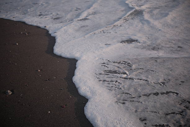 Waves gently crash onto a sandy shoreline under the warm glow of a setting sun. The orange and golden hues reflect off the water, creating a serene, picturesque scene. The beach is devoid of people, emphasizing the tranquility and natural beauty of the moment.