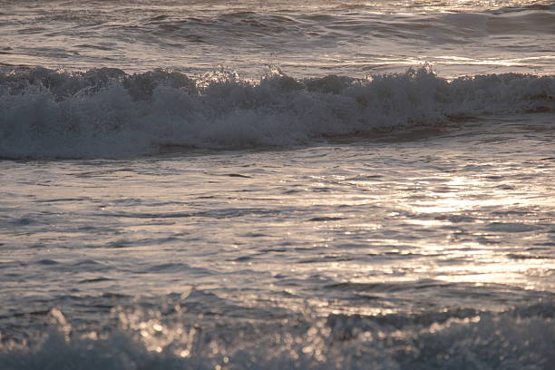 Waves gently crash onto a sandy shoreline under the warm glow of a setting sun. The orange and golden hues reflect off the water, creating a serene, picturesque scene. The beach is devoid of people, emphasizing the tranquility and natural beauty of the moment.