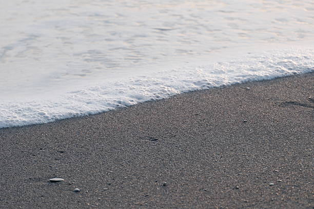 Waves gently crash onto a sandy shoreline under the warm glow of a setting sun. The orange and golden hues reflect off the water, creating a serene, picturesque scene. The beach is devoid of people, emphasizing the tranquility and natural beauty of the moment.